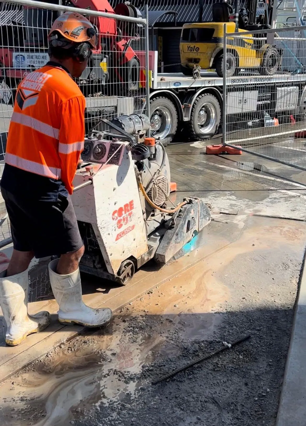 Direct Concrete operator running a road saw inside temporary fencing on a Timaru commercial site, truck and telehandler behind